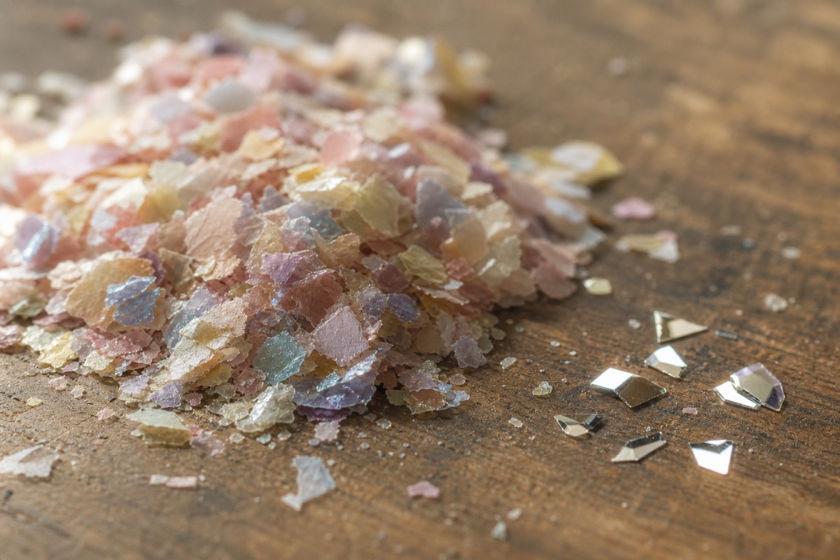 Nahaufnahme eines Häufchens matteren Zellulose-Bio-Glitters neben einigen glänzenden synthetischen PET-Partikeln auf einer Holzoberfläche.