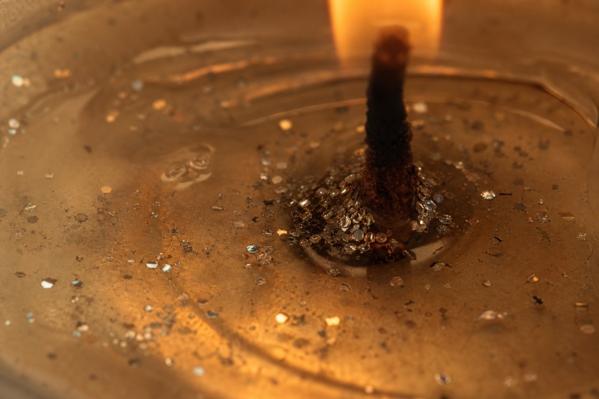 Makroaufnahme von geschmolzenem durchsichtigem Wachs am Docht einer brennenden Kerze, in dem winzige silberne Glitzerpartikel absinken.