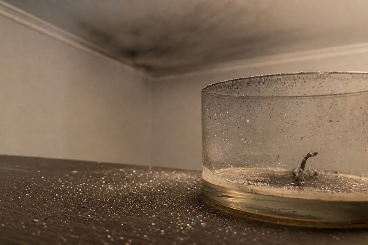 Winzige Glitzerpartikel über den Tisch verstreut rund um eine heruntergebrannte Kerze, im Hintergrund ein dünner grauer Rußfilm an der Decke.